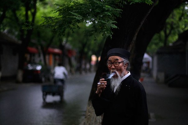 Китаец в традиционном костюме курит сигарету возле своего магазина, на аллее в Пекине. 