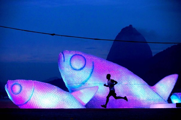 Человек совершает пробежку рано утром на пляже Ботафого у огромных скульптур из пластиковых бутылок, Рио-де-Жанейро, Бразилия. На фоне — гора Сахарная голова. 